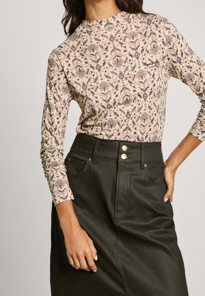 Weibliches Model trägt ein enganliegendes langärmeliges cremefarbenes Oberteil mit Blumenmustern, kombiniert mit einem hoch taillierten dunkelgrünen Rock mit doppelten goldenen Knöpfen.