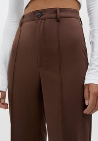 Detailní záběr osoby v hnědých kalhotách s vysokým pasem a zapínáním na knoflík, s bílou dlouhým rukávem nahoře, ruce uvolněně podél těla.