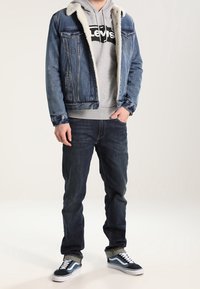 Hombre con chaqueta de mezclilla azul con forro de forro polar blanco, sudadera gris de Levi's, jeans oscuros y zapatillas en blanco y negro, de pie con una mano en el bolsillo.
