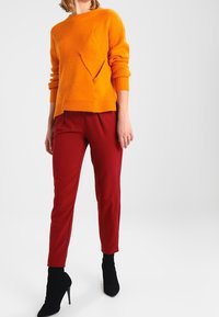 Orange stickad tröja med lång ärm och ett texturerat mönster, kombinerad med röda skräddarsydda byxor och svarta ankelstövlar.