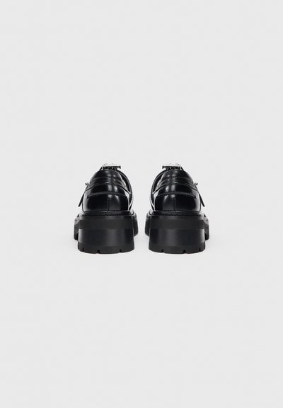 Un paio di scarpe platform in pelle nera lucidate con suole spesse, viste da dietro su sfondo bianco.