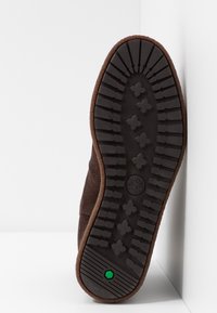 Suela de zapato de ante marrón con una suela de goma negra texturizada, que presenta un logo hexagonal verde y patrones de profundidad para un mejor agarre.