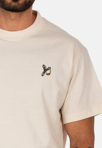 T-shirt in cotone beige a maniche corte con un piccolo design ricamato di attrezzi in marrone, grigio e blu nella zona sinistra del petto.