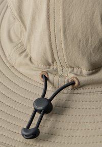 Κοντινό πλάνο μπεζ υφασμάτινου καπέλου με μαύρο ρυθμιζόμενο κορδόνι και πλαστικό κούμπωμα περασμένο μέσα από ραμμένα ρουμπά.