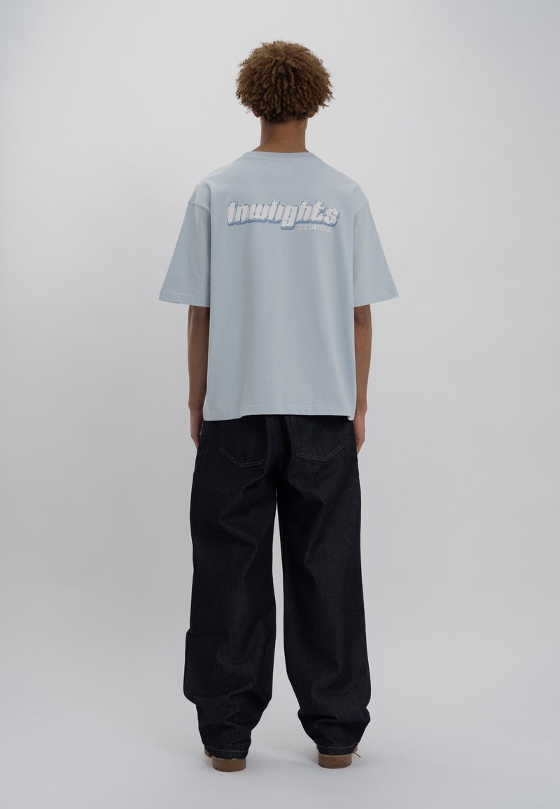 Personne avec des cheveux bouclés portant un t-shirt oversized bleu clair avec le texte "lowlights studios", un jean large sombre et des chaussures marron, de dos.