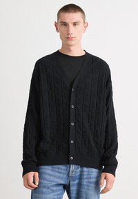 Mustakudottu cardigan, jossa on timanttimäinen kuvio, v-kaulus, nappikiinnitys ja resorit. Yhdistetty vaaleansinisiin farkkuihin.