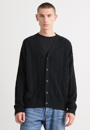 Melnā adīta kardigāna ar dimanta rakstu, ar V veida kakla izgriezumu, pogu aizdari un ribotiem piedurknēm. Saskaņots ar gaiši ziliem džinsiem.