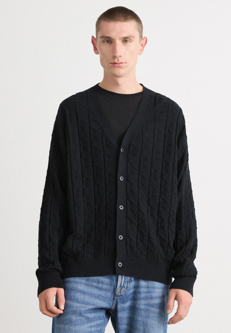 Mustakudottu cardigan, jossa on timanttimäinen kuvio, v-kaulus, nappikiinnitys ja resorit. Yhdistetty vaaleansinisiin farkkuihin.