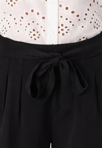 Pantaloni neri in tessuto con cintura legata, pieghe e texture liscia, abbinati a una maglietta in broderie anglaise bianca.