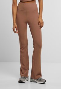 Bruine high-waisted flared legging gemaakt van rekbaar materiaal met een gladde textuur. Draagt met grijze sportschoenen.