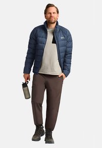 Gepolsterte marineblaue Jacke mit hohem Kragen, grauem Hemd und brauner Hose. Schwarze Wanderschuhe und graue Wasserflasche. Lässige Outdoor-Bekleidung.
