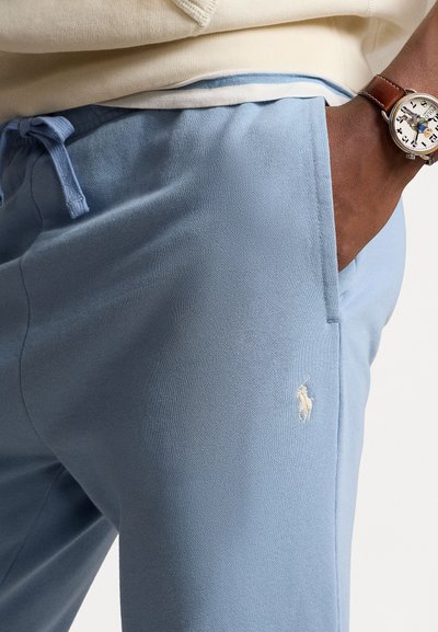 Lyseblå joggebukser laget av mykt stoff, med elastisk midjebånd og snor, samt en liten hvit brodert logo på låret.