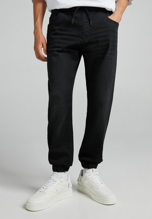 Man met zwarte joggingbroek en witte sneakers staat op een effen achtergrond met handen in de zakken.