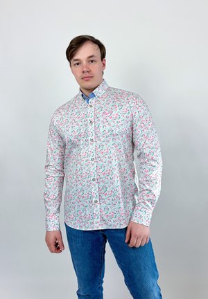 Jeune homme debout, portant une chemise blanche à manches longues avec un motif floral rose et vert et un jean bleu sur un fond uni.