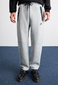 Pantaloni de trening gri cu buzunare cu fermoar negre, talie ajustabilă și logo-ul Nike. Purtați cu pantofi negri cu un finisaj texturat.