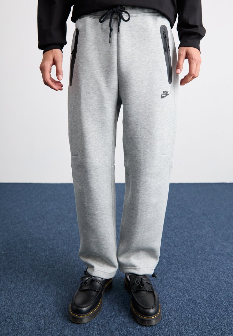 Pelēki sporta bikses ar melniem rāvējslēdzēja kabatām, regulējamu jostu un Nike logotipu. Valkātas ar melnām kurpēm ar teksturētu virsmu.