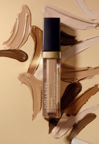 Klar slange med svart og gull hette, som inneholder en lys brun concealer. Rundt er det virvler av concealer i forskjellige nyanser på en beige bakgrunn.