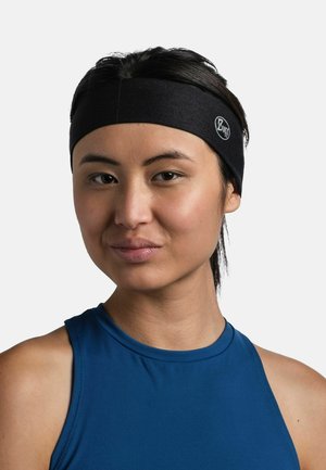 Joven mujer con cabello oscuro que lleva una cinta negra Buff y una camiseta azul sin mangas, mirando hacia adelante con expresión neutral.