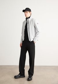 Ανοιχτό γκρι μπουφάν με υψηλό γιακά, μαύρο φερμουάρ και λογότ�υπο. Φορεμένο πάνω από μια μαύρη μπλούζα και παντελόνι, συνδυασμένο με μαύρα παπούτσια και καπέλο.