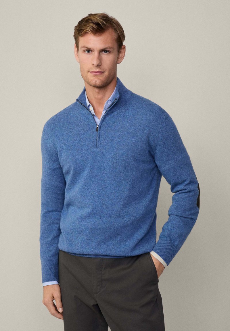 Maglione azzurro con zip fino a metà realizzato in tessuto morbido, con maniche lunghe e toppe nere sui gomiti, abbinato a una camicia colletto blu chiaro sotto.