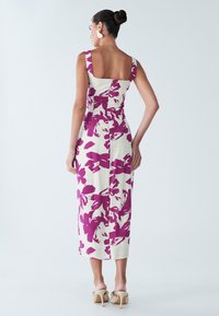 Robe ajustée longueur genoux en crème avec un motif floral violet audacieux. Encolure carrée, larges bretelles et détails de couture au dos. Sandales à talons.