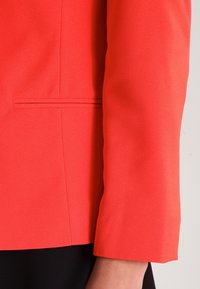 Ljusa korallblazer i slät textur, skräddarsydd passform och långa ärmar. Har en enkel dekorativ söm nära fickområdet.