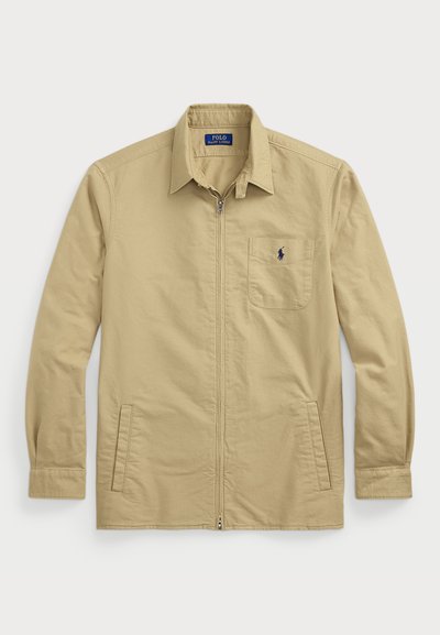 Gaišs bezrūpīgs jaku ar garām piedurknēm, punktveida apkakli, priekšējām kabatām un mazu izšūtu logotipu virs kreisās kabatas. Kokvilnas materiāls.