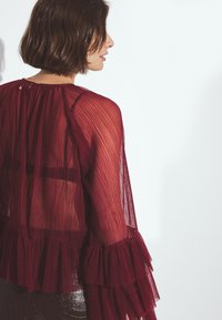 Bordeaux plissé blouse met gerimpelde mouwen, doorschijnende stof en een ronde halslijn. Textuurdetail op de mouwen en achterkant benadrukt het ontwerp.