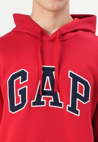 Rode katoenen hoodie met een vooraanzicht kangoeroezak, voorzien van een groot marineblauw "GAP"-logo omrand met wit. Verstelbare trekkoordkap.