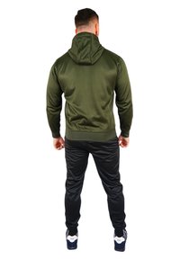 Felpa verde oliva con cappuccio dalla trama liscia, abbinata a pantaloni neri a vita alta. L'outfit presenta polsini elasticizzati e un design minimalista.