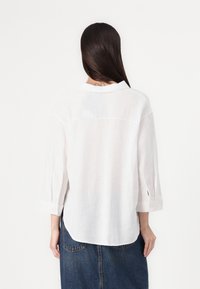 Camicia bianca di lino con colletto, maniche a tre quarti e dettagli in button. Presenta un orlo leggermente curvo e una texture leggera.