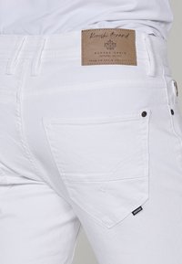 Pogled od blizu na osebo, ki nosi bele denim hlače in prikazuje zadnji žep ter rjavo usnjeno nalepko blagovne znamke z napisom "Rugged Denim".