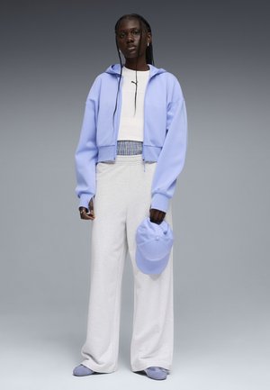 Personne grande avec de longues tresses portant un sweat à capuche violet clair zippé, un haut court blanc, un pantalon blanc ample, et tenant une casquette violette.