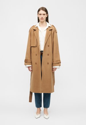 Vrouw draagt een lange dubbele rij knopen camelkleurige jas over een wit overhemd, blauwe jeans en witte puntige schoenen, staand tegen een effen achtergrond.