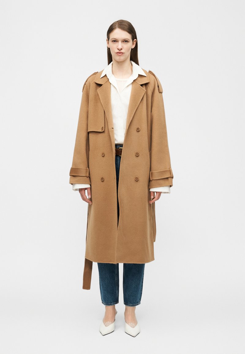 Γυναίκα που φοράει μακριά καμηλοπάρδαλη διπλόκλειστο παλτό πάνω από λευκό πουκάμισο, μπλε τζιν και λευκά μυτερά παπούτσια, όρθια μπροστά σε ένα απλό φόντο.