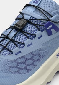 Αθλητικό παπούτσι ανοιχτού γαλάζιου χρώματος με υφή στον άνω μέρος, που φέρει μοτίβο από εξαγωνικά κελιά, ελαστικά κορδόνια και μοβ λεπτομέρειες, με λευκή σόλα.