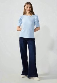 Lichtblauwe getextureerde top met puffmouwen, gecombineerd met donkere flare jeans en witte sneakers. Eenvoudig ontwerp, zonder zichtbare patronen of accenten.