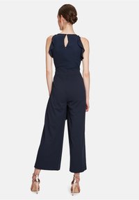 Tengerészkék overál, fodros pántokkal és kulcslyuk alakú hátrészletel. Széles lábakkal és formás derékkal rendelkezik, puha, rugalmas anyagból készült.