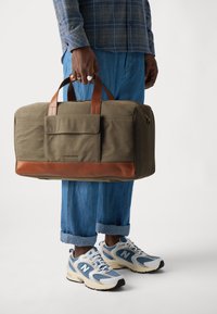 Olive green bag with brown leather accents and handles; front flap pocket; soft fabric; held in hand, worn with light blue cuffed trousers and sneakers.