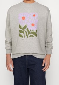 Felpa grigia chiara con grafica floreale rosa e arancione e foglie verdi, abbinata a jeans blu scuro, indossata da una persona in piedi.