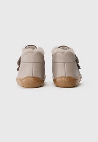 Een paar beige leren peuterschoenen met een bruine rubberen zool en beige fleece voering, gezien vanaf de achterkant op een witte achtergrond.