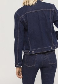 Mörkblå denimjacka med en figurnära design, synlig sömnad och metallaccenter. Har knäppningar med knappar och framfickor.
