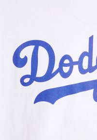 Bílý bavlněný tričko s velkým modrým kurzivním textem "Dodgers." Materiál má hladkou texturu bez zjevných vzorů.