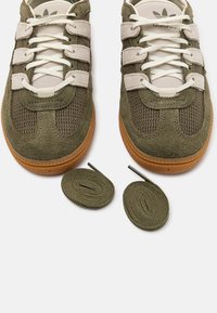 Pár sneakers z olivově zelené semiše a síťoviny s bílými tkaničkami a gumovými podrážkami, zobrazený s dvěma extra olivově zelenými tkaničkami níže.