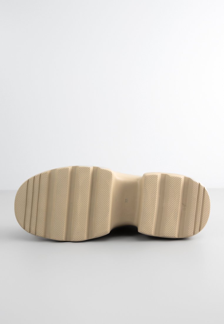 Sohle eines beigen Schuhs mit breiten horizontalen Rillen und strukturierten Abschnitten, vor einem schlichten hellen Hintergrund gezeigt.
