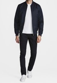 Chaqueta bomber azul marino con cuello acanalado y cierre de cremallera, combinada con camiseta blanca y vaqueros oscuros de corte ajustado, rematada con zapatillas blancas.