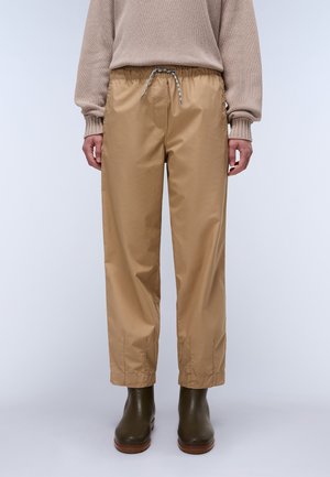 Personne portant un pantalon beige à cordon, un pull en maille beige et des bottines vert foncé, debout devant un fond clair uni.