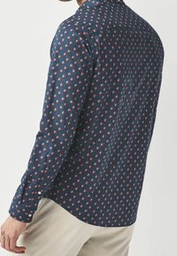 Man gekleed in een marineblauwe lange mouwen shirt met een klein rood en wit bloemenpatroon en lichtbeige broeken, getoond van achter- en zijdeaanzicht.