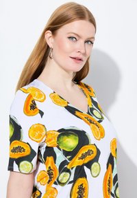Camiseta blanca de cuello en V con un estampado colorido de frutas, incluyendo patrones de papaya, lima y naranja; tela suave con mangas cortas.