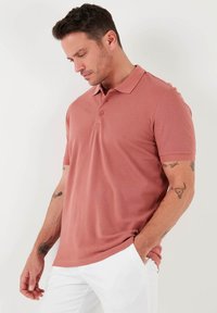 Polo särk tolmuroosa värviga, valmistatud tekstureeritud kangast. Omab klassikalist krae, kolme nupuga kinnitusvärava ja lühikesi varrukaid.